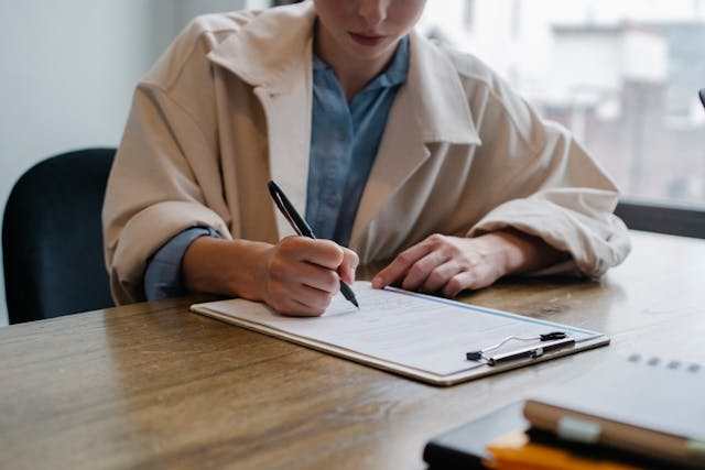 person-sitting-at-a-table-writing-on-a-piece-of-paper-clipped-on-a-clipboard